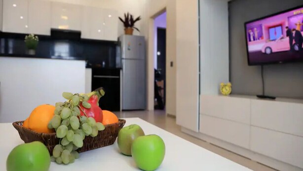 A modern kitchen with white cabinets, a refrigerator, and a television displaying a show. A fruit basket with grapes, apples, and an orange is on the counter. - Kristal Absheron 110 (Baku)