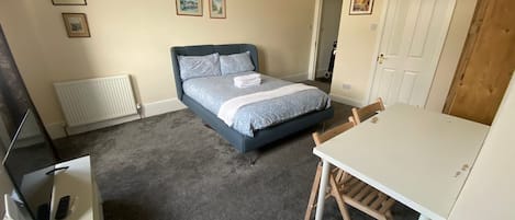A bedroom with a bed, a desk with a chair, a radiator, and framed pictures on the wall.
