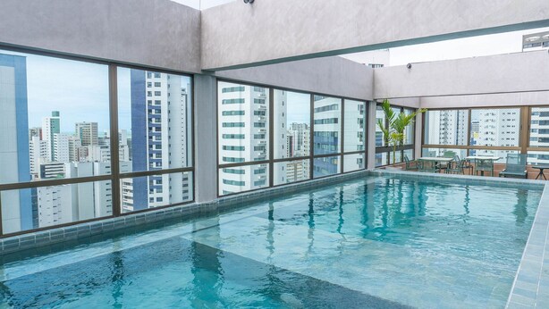 An indoor swimming pool with large windows offering city views. - Flats no Rooftop 470 por Yolo (Recife)