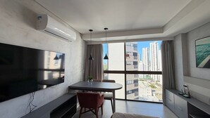 A modern living room with a flat-screen TV, a dining table, and a large window offering a city view. - Flats no Rooftop 470 por Yolo (Recife)