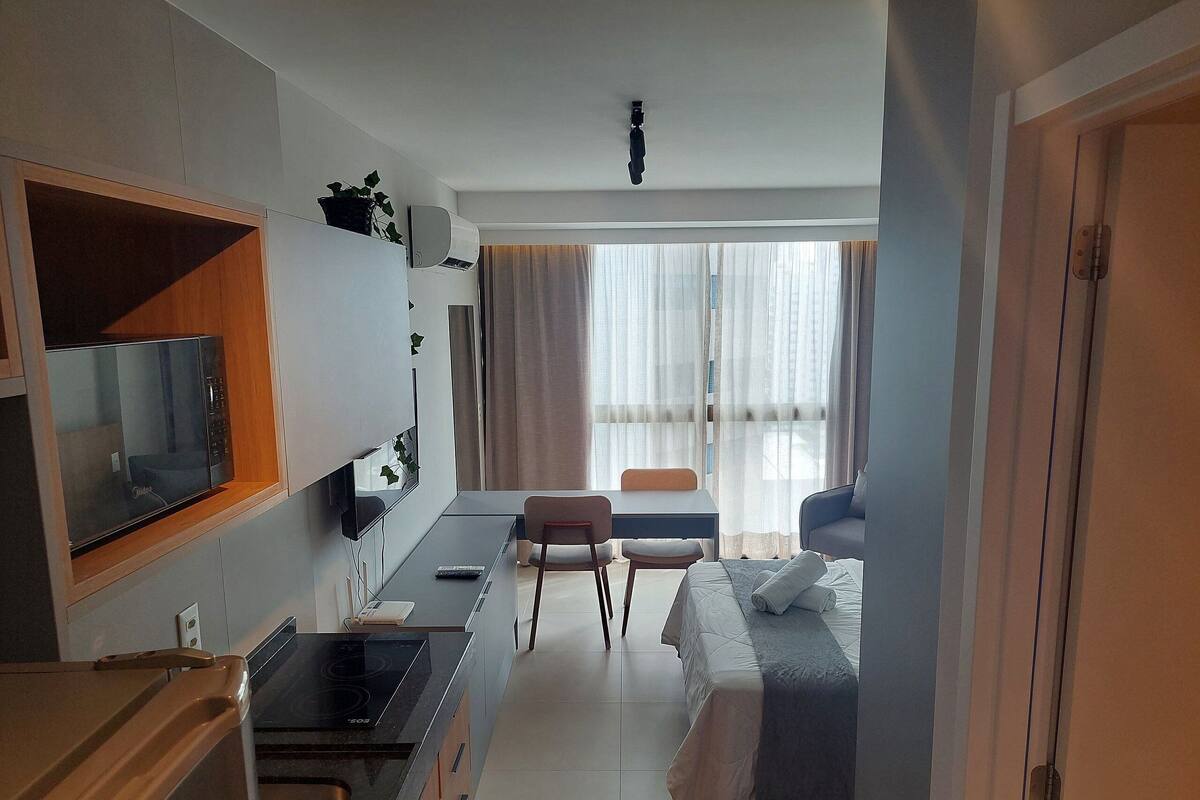 A compact hotel room with a kitchenette, a bed, a seating area, and a large window.