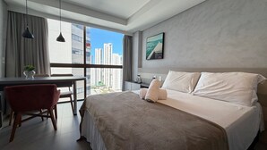 A modern hotel room with a large bed, a desk, a chair, and a view of tall buildings. - Flats no Rooftop 470 por Yolo (Recife)