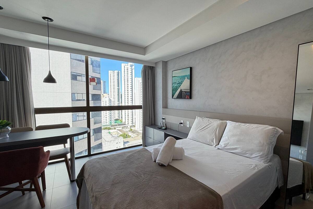 A modern hotel room with a large bed, a desk, and a view of high-rise buildings.