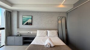 A modern hotel room with a bed, a desk, a chair, a large window, and a painting on the wall. - Flats no Rooftop 470 por Yolo (Recife)