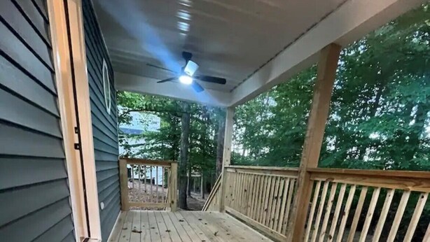 Una terraza de madera con barandilla, un ventilador de techo y vista a árboles al fondo.