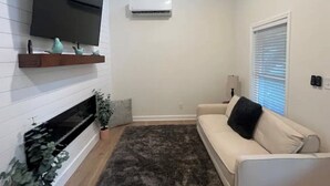 Una sala de estar con un televisor de pantalla plana montado en la pared, un aire acondicionado blanco, un sofá beige, un tapete oscuro y una pequeña planta.