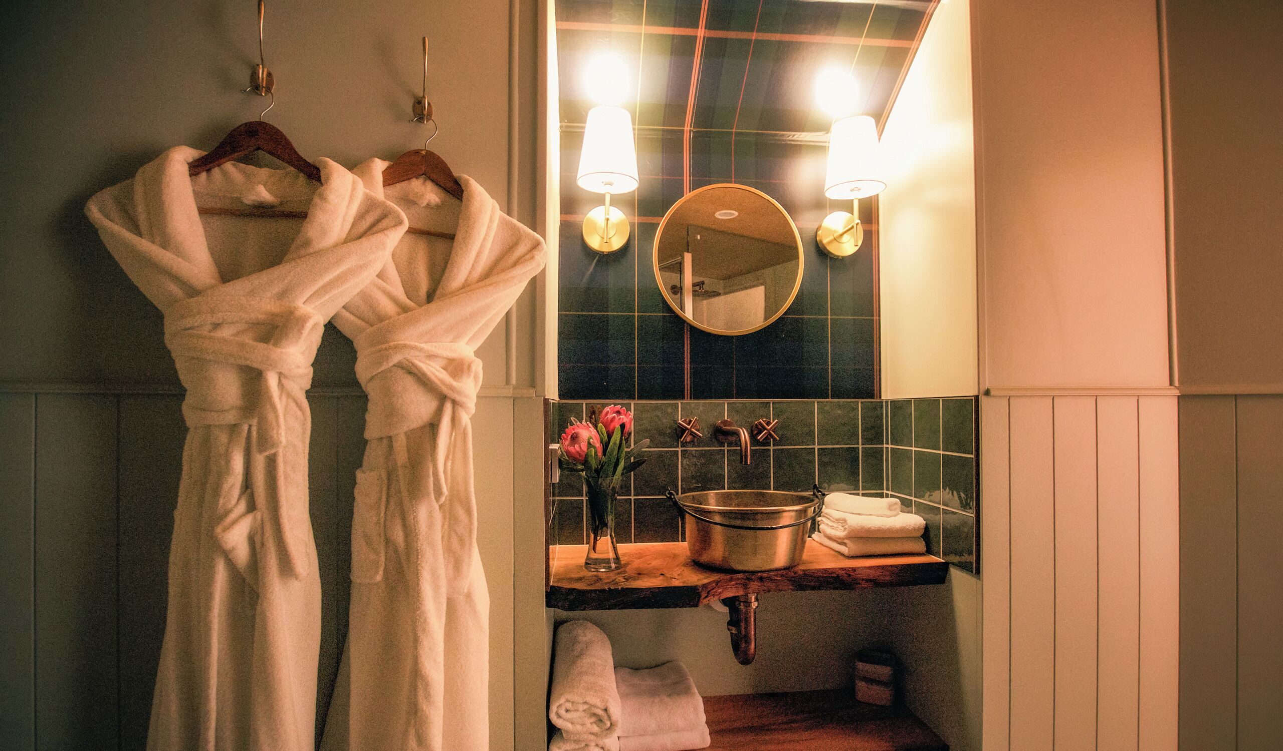 Une salle de bain avec deux peignoirs blancs suspendus à un porte-serviettes, un miroir rond, un lavabo avec un pot et deux appliques murales.