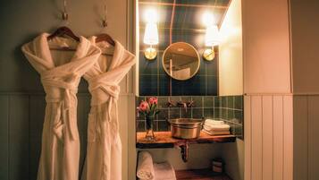 Une salle de bain avec deux peignoirs blancs suspendus à un porte-serviettes, un miroir rond, un lavabo avec un pot et deux appliques murales.