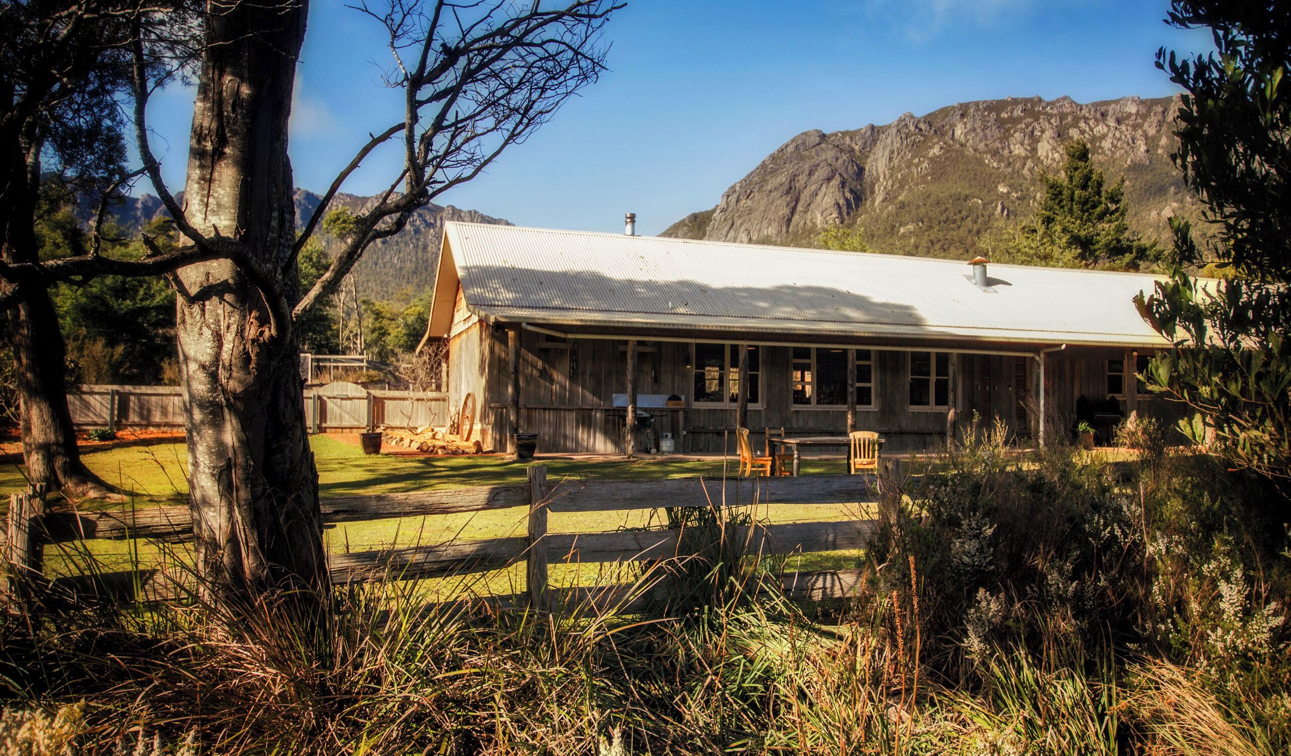 A rustic wooden building with a corrugated metal roof, surrounded by a fenced yard and trees, with mountains in the background.