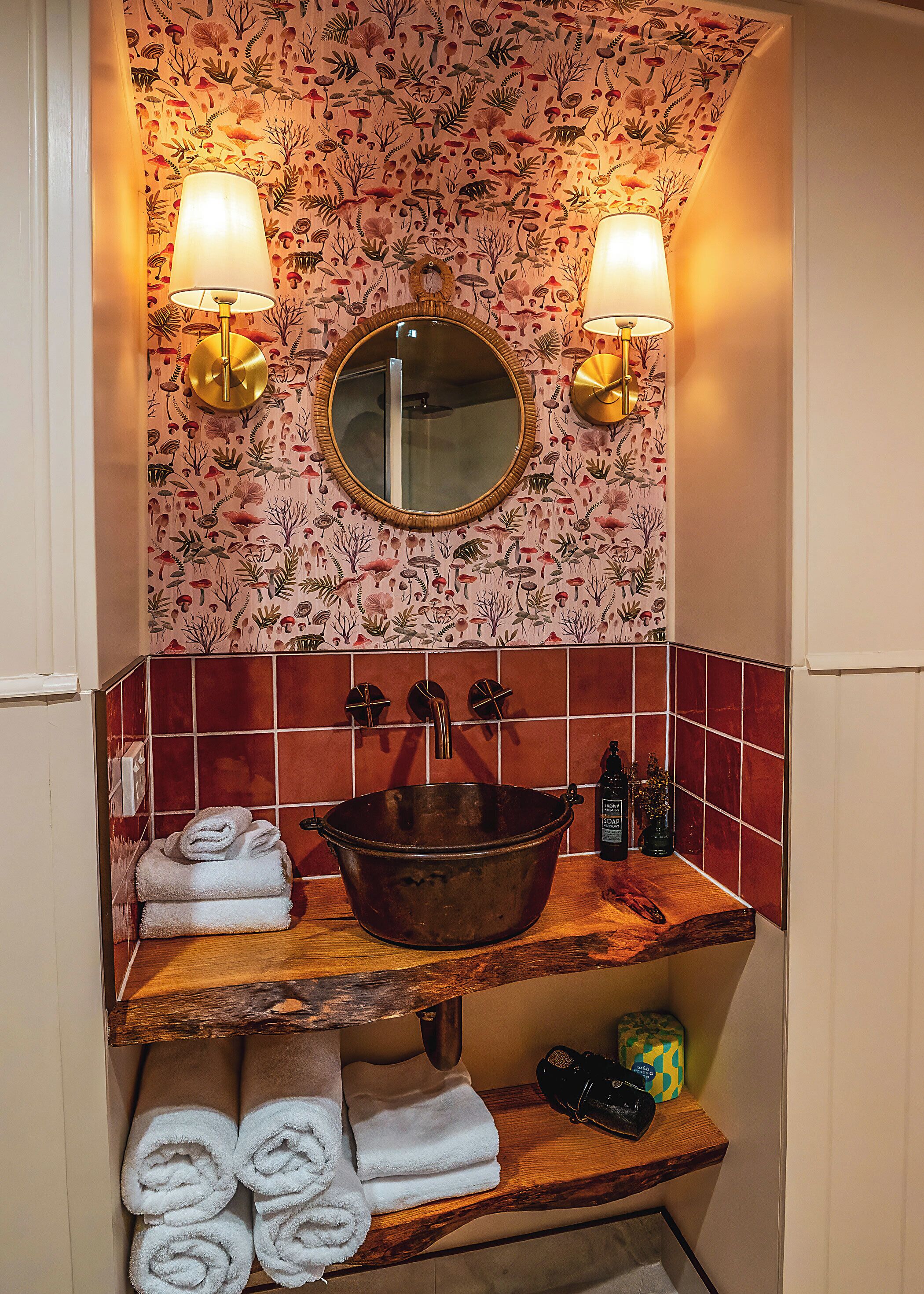 Une salle de bain avec un meuble-lavabo en bois, un miroir rond, deux lampes fixées au mur, un grand lavabo et un porte-serviettes avec des serviettes pliées.