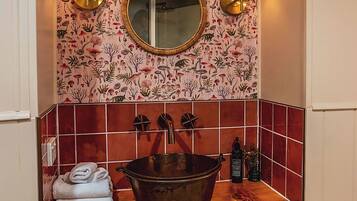 Une salle de bain avec un meuble-lavabo en bois, un miroir rond, deux lampes fixées au mur, un grand lavabo et un porte-serviettes avec des serviettes pliées.