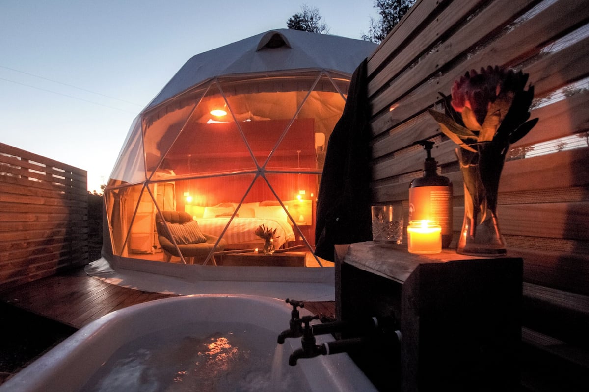 Une structure en forme de dôme géodésique comprenant un lit et une petite table avec une bougie, le tout entouré de murs en bois et d’une terrasse en bois.