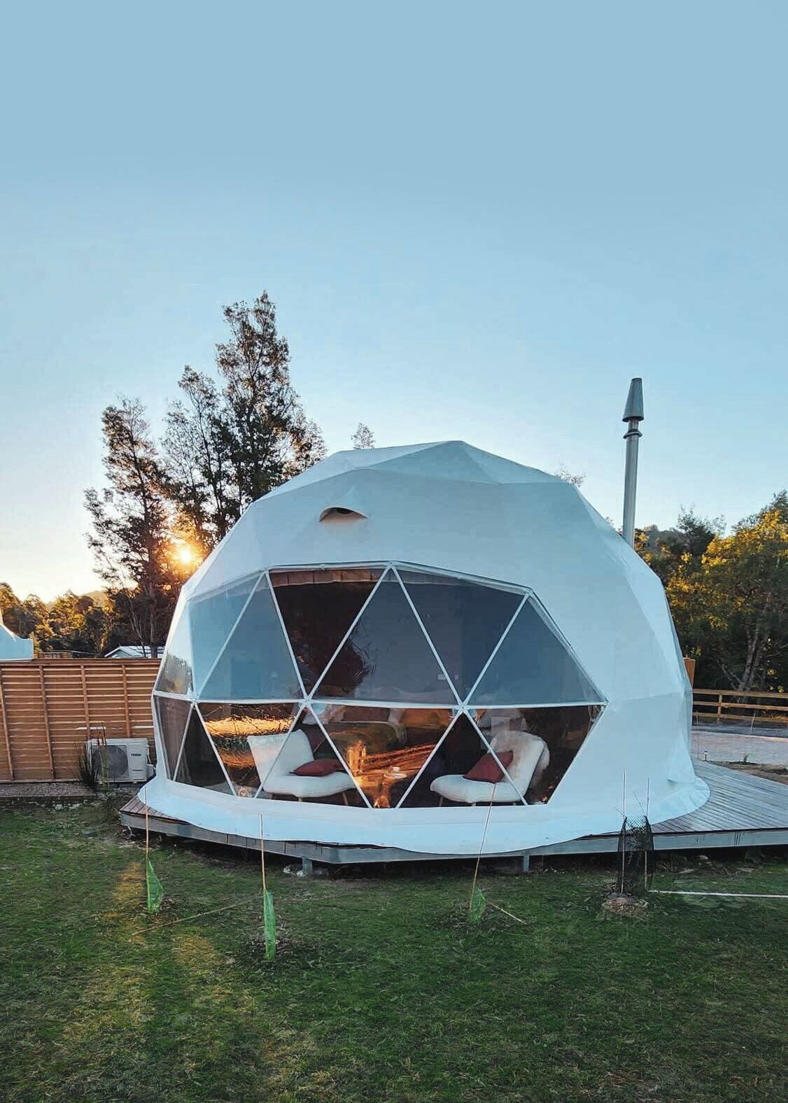 Una estructura de cúpula geodésica con un brasero en su interior, rodeada por una terraza de madera y un área de césped.