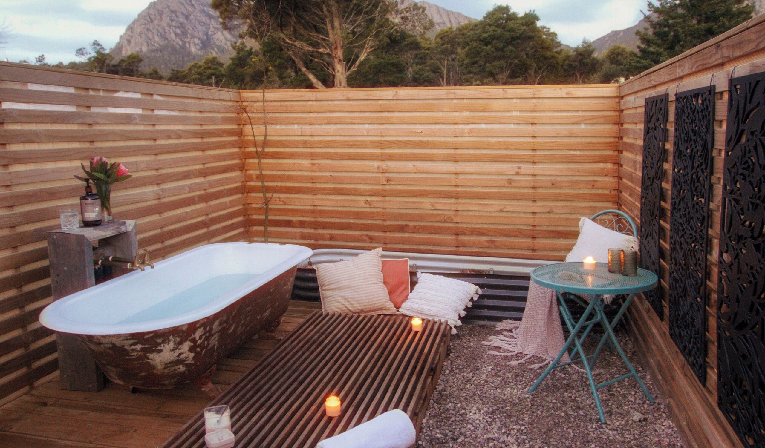 Un área de spa al aire libre con cerramiento de madera, una bañera antigua, una camilla de masaje y una mesa pequeña con velas.