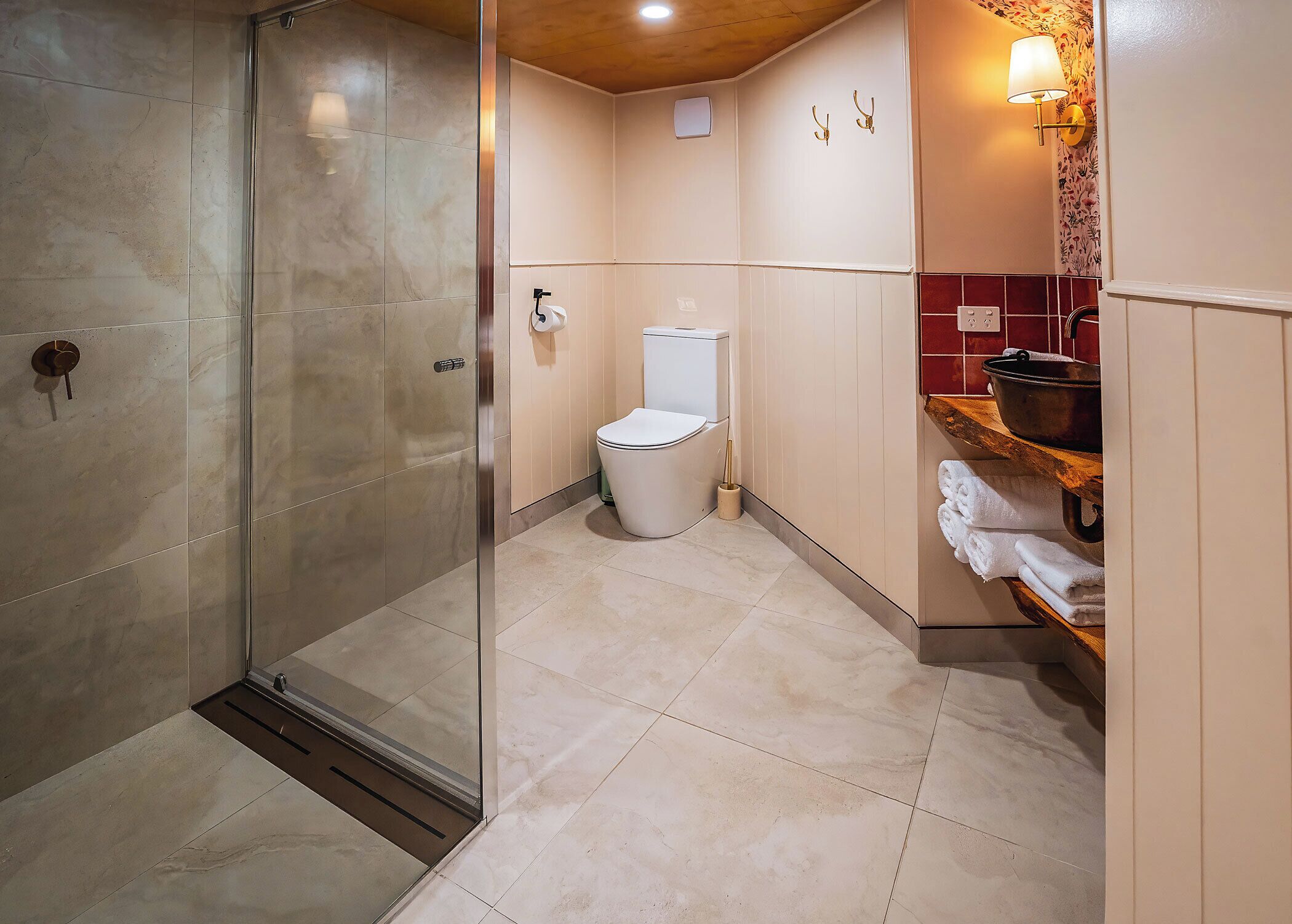 A modern bathroom with a glass shower enclosure, a toilet, and a sink area with a wooden shelf.