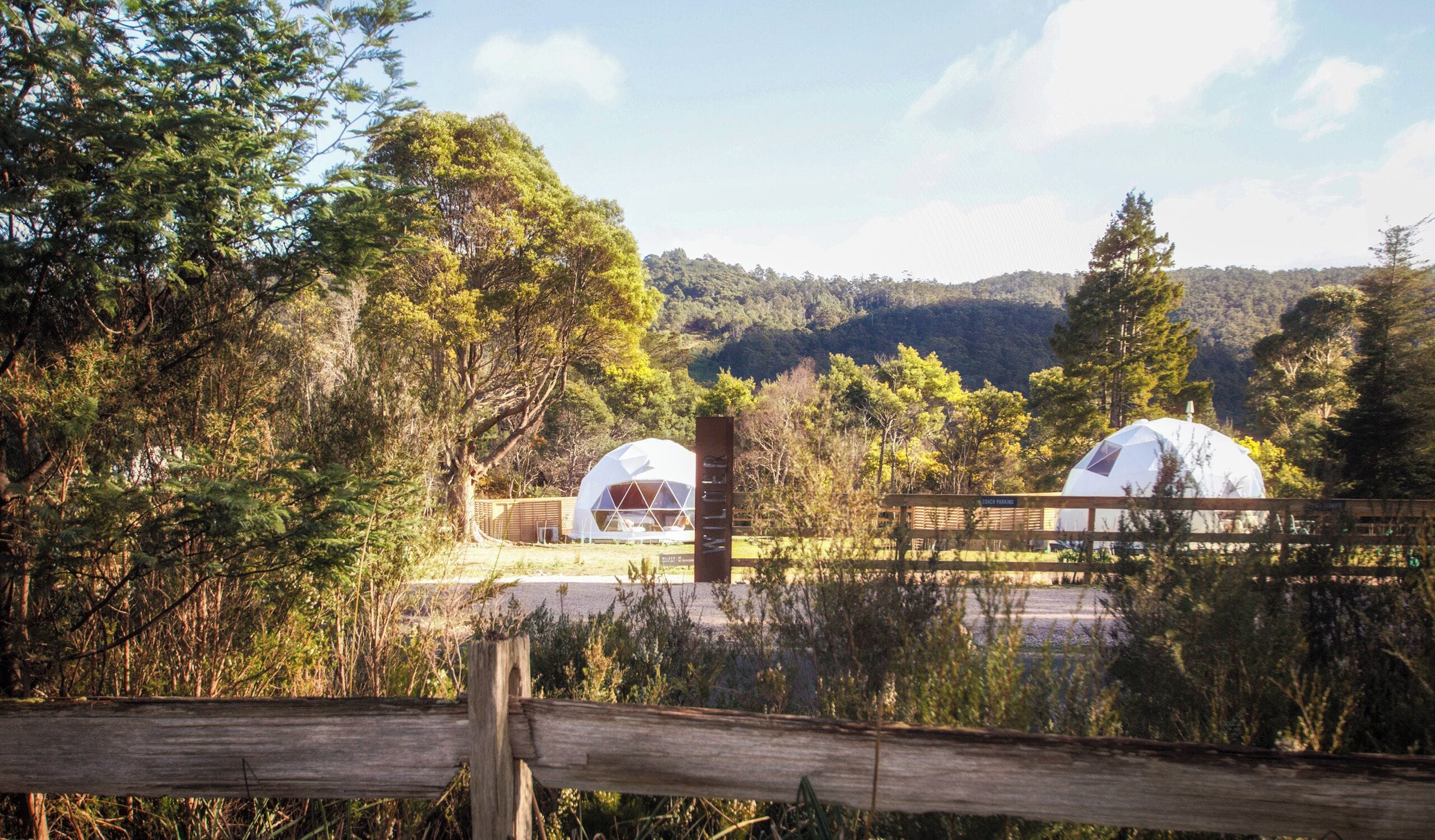 Two geodesic dome structures in a forested area with a wooden fence in the foreground.