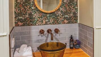 A bathroom with a wooden vanity, a brass basin, two wall-mounted lamps, a mirror, and a tiled backsplash.