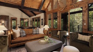 A cozy living room with a brown leather sofa, a grey ottoman, and a brown armchair. There are multiple windows providing a view of the outdoors. - Iron Tree Lodge Blue Ridge GA (Blue Ridge)