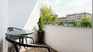 Un balcón con dos sillones de mimbre y una mesita, una planta en maceta y vistas de edificios al otro lado de un muro blanco.