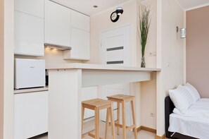 Een kleine kamer met een kitchenette, een bed en een klein houten krukje.