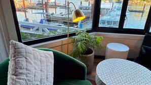 A room with a green sofa, a white pillow, a small round table, and a view of a marina with boats and mountains.