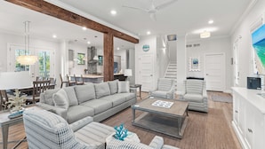 A spacious living room with a gray sofa, patterned armchairs, and a coffee table. A kitchen is visible in the background with wooden beams and white cabinets. - Bike, EV Charger, New Rental! Golf Cart Included, Pool, 30A (Santa Rosa Beach)