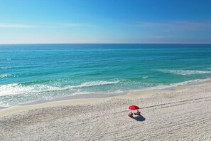 A beach with a red umbrella and two people sitting on chairs.