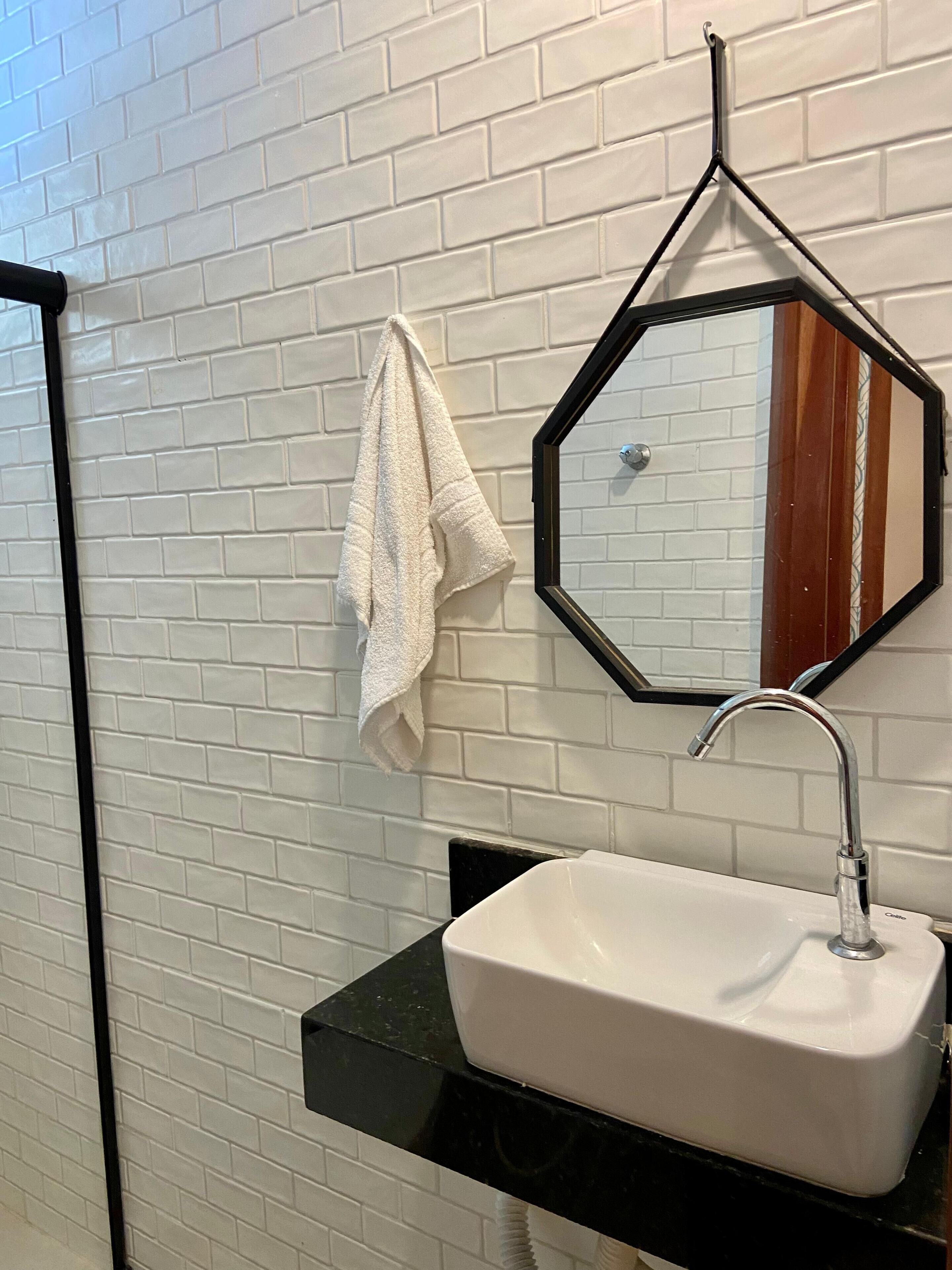 Une salle de bain avec un mur recouvert de carreaux blancs, un plan de toilette noir, un lavabo blanc, un robinet chromé, une serviette accrochée au mur et un miroir hexagonal.