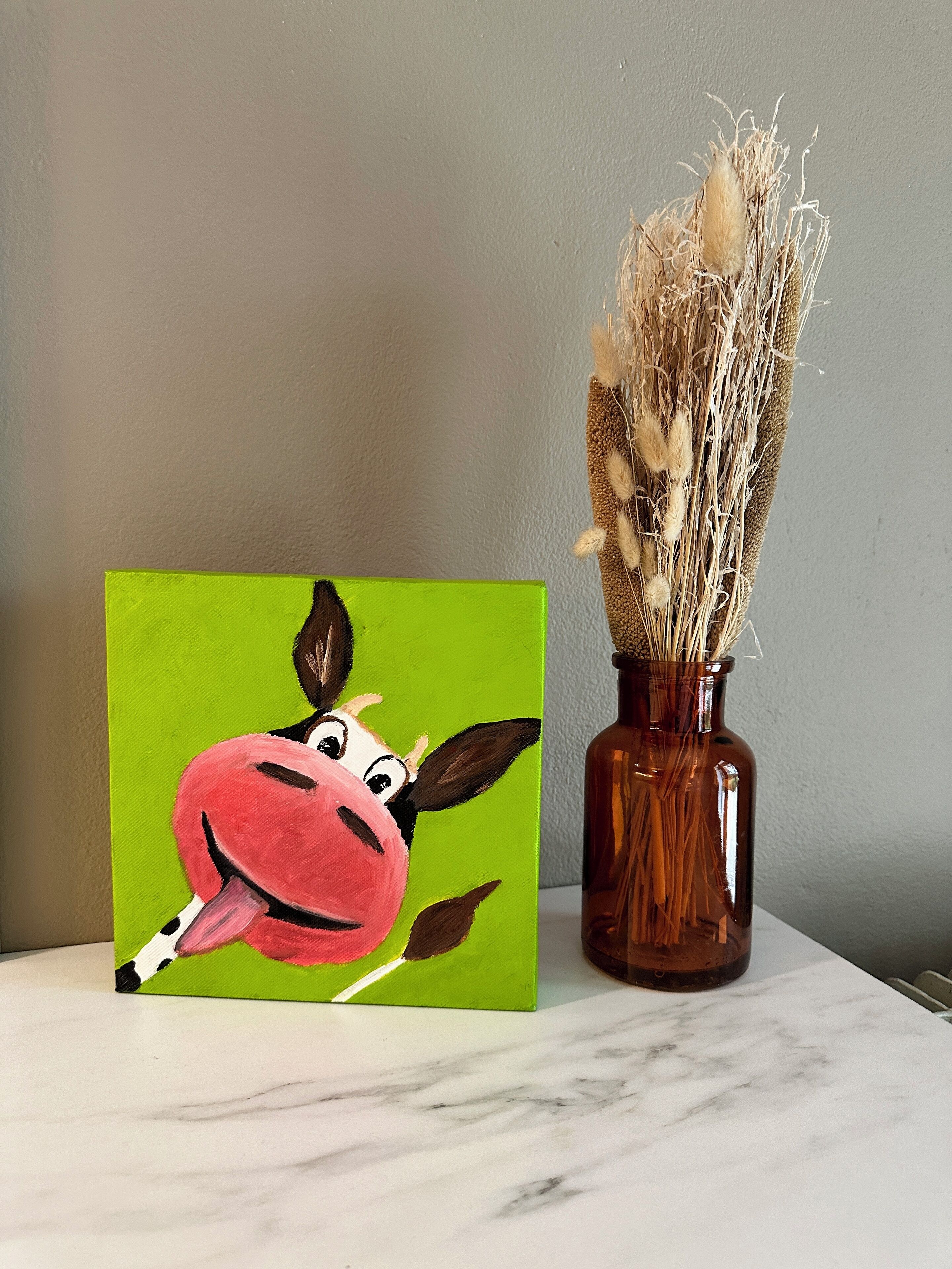 A painting of a cartoon cow on a green background next to a vase with dried plants.