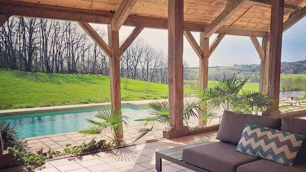 A covered outdoor seating area with a pool, wooden beams, and a view of a green landscape. - Luxury Holiday Home Paulhiac - Large Pool (Paulhiac)