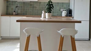 A modern kitchen with a central island, two bar stools, a hanging light fixture, a refrigerator, and a tiled backsplash. - Le Champ du Roy (Chasse-sur-Rhône)