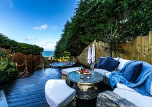 Relax on the deck with a hot tub, cushioned sofa, and round tables amidst lush greenery.