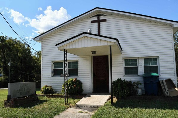Uma igreja branca com uma cruz na fachada, um alpendre e uma placa com a inscrição 'First Apostolic Church'.