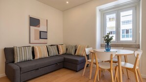 A modern living room with a grey sectional sofa, striped cushions, a round wooden table with chairs, and a large window with a view of buildings.