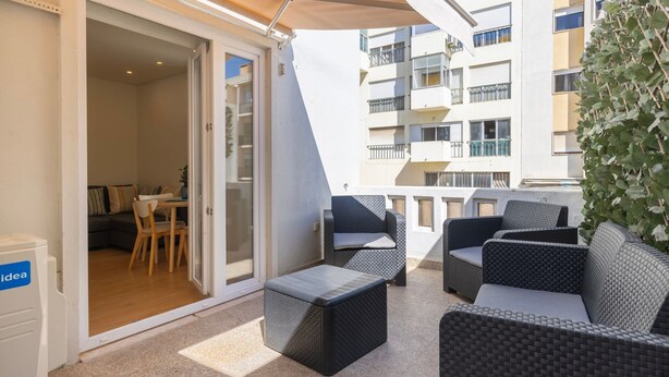 An outdoor balcony with wicker furniture, a dining area, and a view of a multi-story building.