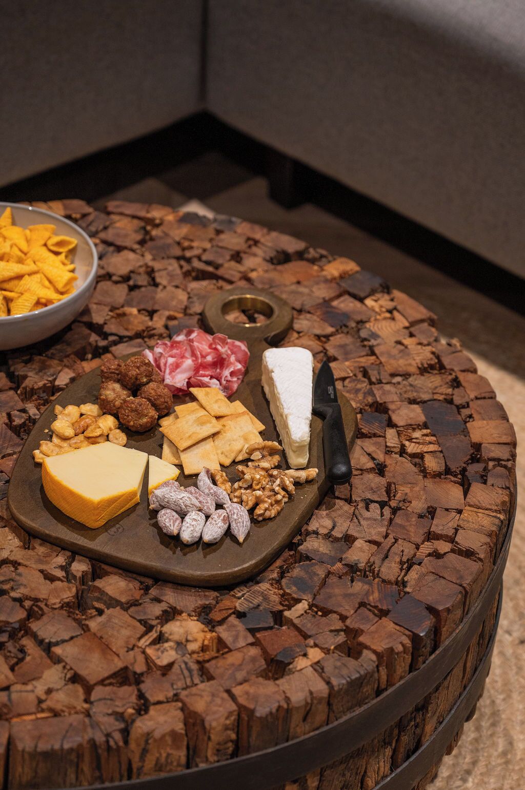 A round wooden table with a cheese board containing various cheeses, nuts, and meats.