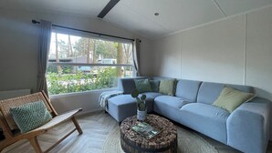 A modern living room with a grey sofa, a round coffee table, a wicker chair, and a large window with a view of the outside. - Forest Finest, luxury chalet with HOT TUB beautiful canopy and beautiful garden (Hoenderloo)