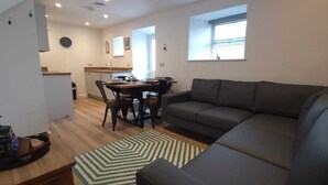A modern living room with a dark grey sofa, a dining table with chairs, and a kitchen area in the background. - Dog Friendly 18th Century Cottage (Saundersfoot)