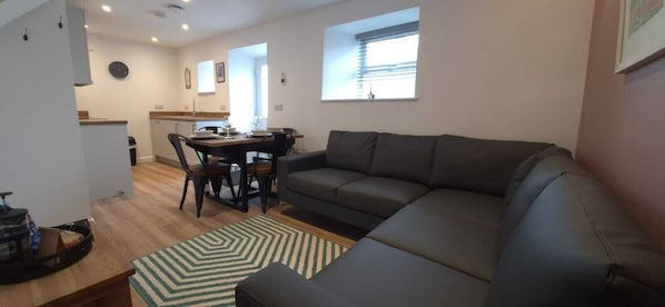 A modern living room with a dark grey sofa, a dining table with chairs, and a kitchen area in the background.