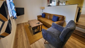 Un salon avec un canapé marron, un fauteuil bleu, une table basse en bois et un téléviseur fixé au mur.