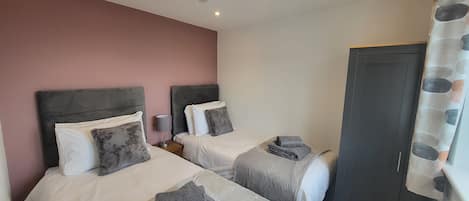 A hotel room with two beds, each with a headboard, a bedside table with a lamp, and a grey towel on the bed.