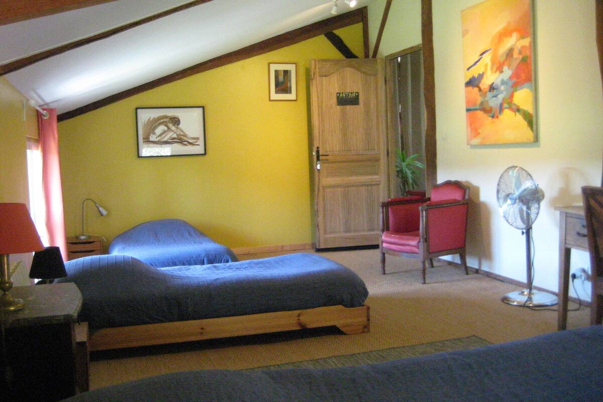 A bedroom with two beds, a wooden door, a red chair, a standing fan, and a painting on the wall.