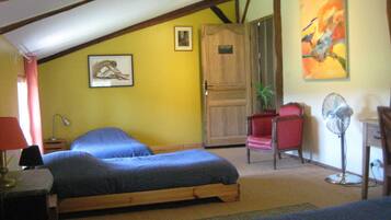 Une chambre avec deux lits, une porte en bois, une chaise rouge, un ventilateur sur pied et un tableau accroché au mur.