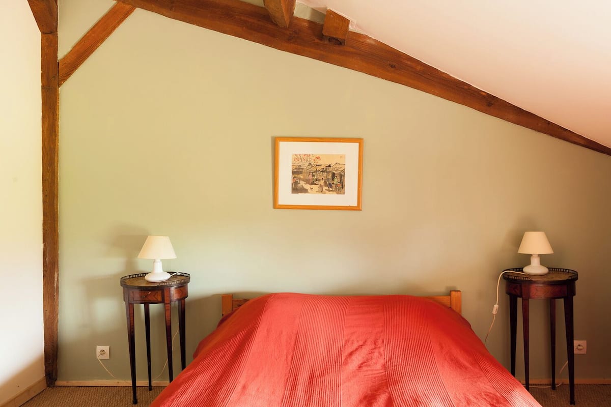 A bedroom with a sloped ceiling, a bed with a red blanket, two bedside tables with lamps, and a framed picture on the wall.
