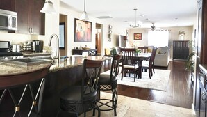 A modern kitchen with dark wooden cabinets, a stainless steel microwave, and a granite countertop. There is a dining area with a table and chairs, and a living room with a sofa and a coffee table. - Charming Craftsman Retreat: 3BR Home w/ Pool & Jacuzzi – Your Perfect Getaway! (Bonita)
