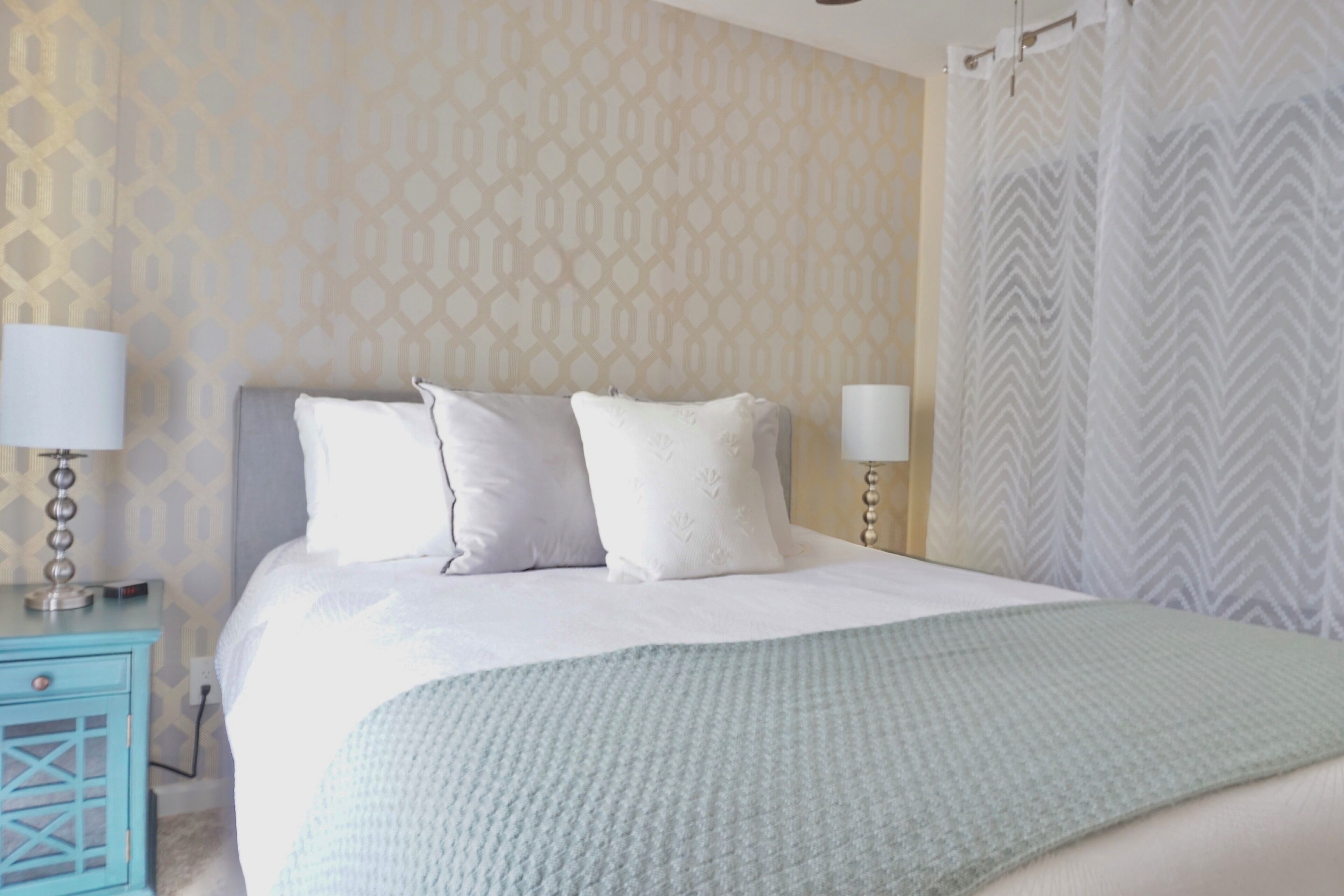 A neatly made bed with a light-colored blanket, two bedside lamps, and a patterned wall.