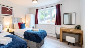 A hotel room with two beds, a desk, a chair, a radiator, a window with curtains, a lamp, and a framed picture on the wall. - Connaught Road House in Aldershot (Aldershot)