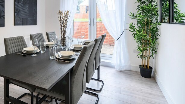 A dining area with a dark wooden table set for six, surrounded by grey upholstered chairs. There are two abstract art pieces on the wall, a potted plant to the right, and a view of a brick wall and greenery outside. - Connaught Road House in Aldershot (Aldershot)