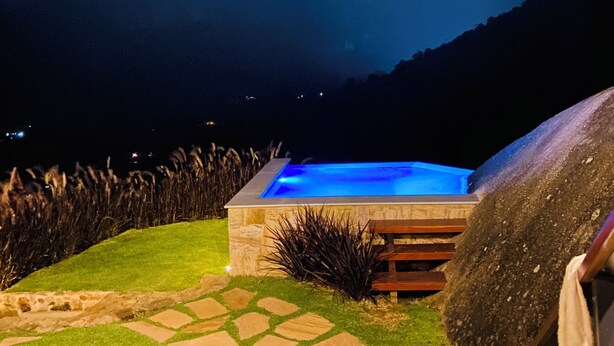 A rectangular pool with a stone border, surrounded by a stone pathway and grass, under a night sky with clouds and a visible moon. - Green Village - Luxury and Comfort you are looking for! (Corrêas Petrópolis)
