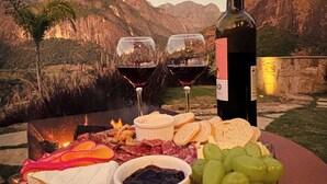 A table set with a bottle of wine, glasses, cheese, bread, grapes, and a small bowl of jam, with a mountainous landscape in the background. - Green Village - Luxury and Comfort you are looking for! (Corrêas Petrópolis)